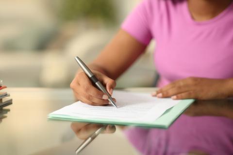 A woman in a pink shirt writing with a pen on a paper.